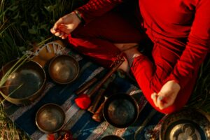 Femme en tenue rouge assise en tailleur dans l'herbe, entourée de bols tibétains en métal, mains en geste mudra, pour bienfaits yoga mental : réduction stress, amélioration sommeil et sérénité
